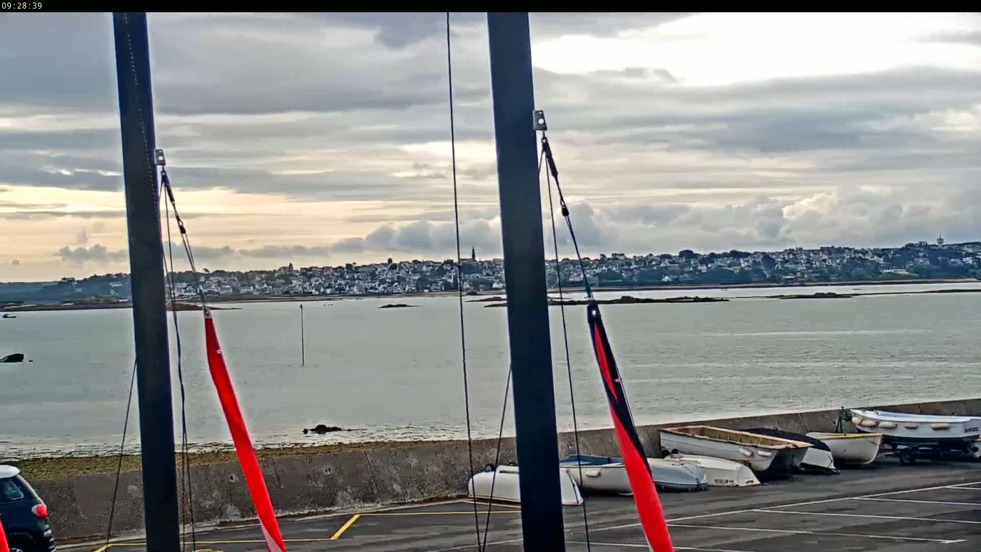 A partly cloudy day overlooks a calm body of water, with several small boats resting on a concrete embankment near a sailboat mast and a distant town visible on the other shore.