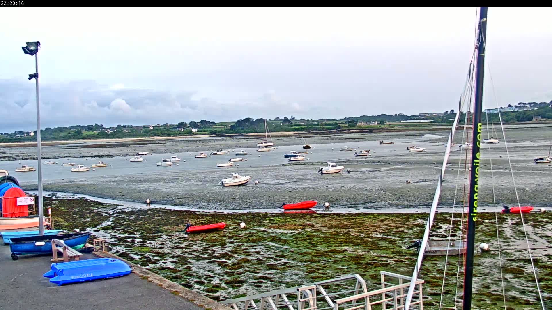 Numerous small boats are moored in a calm bay at low tide, under a partly cloudy sky.