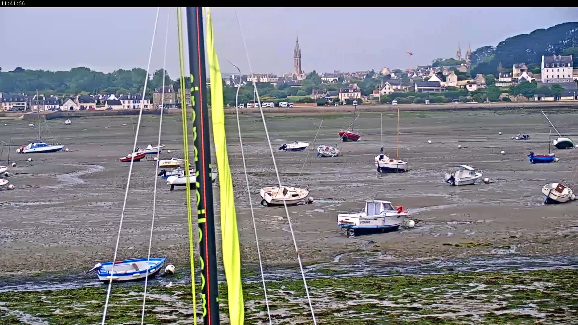 Numerous small boats are grounded on a wide expanse of exposed mudflats, with a town and church visible in the background under an overcast sky.