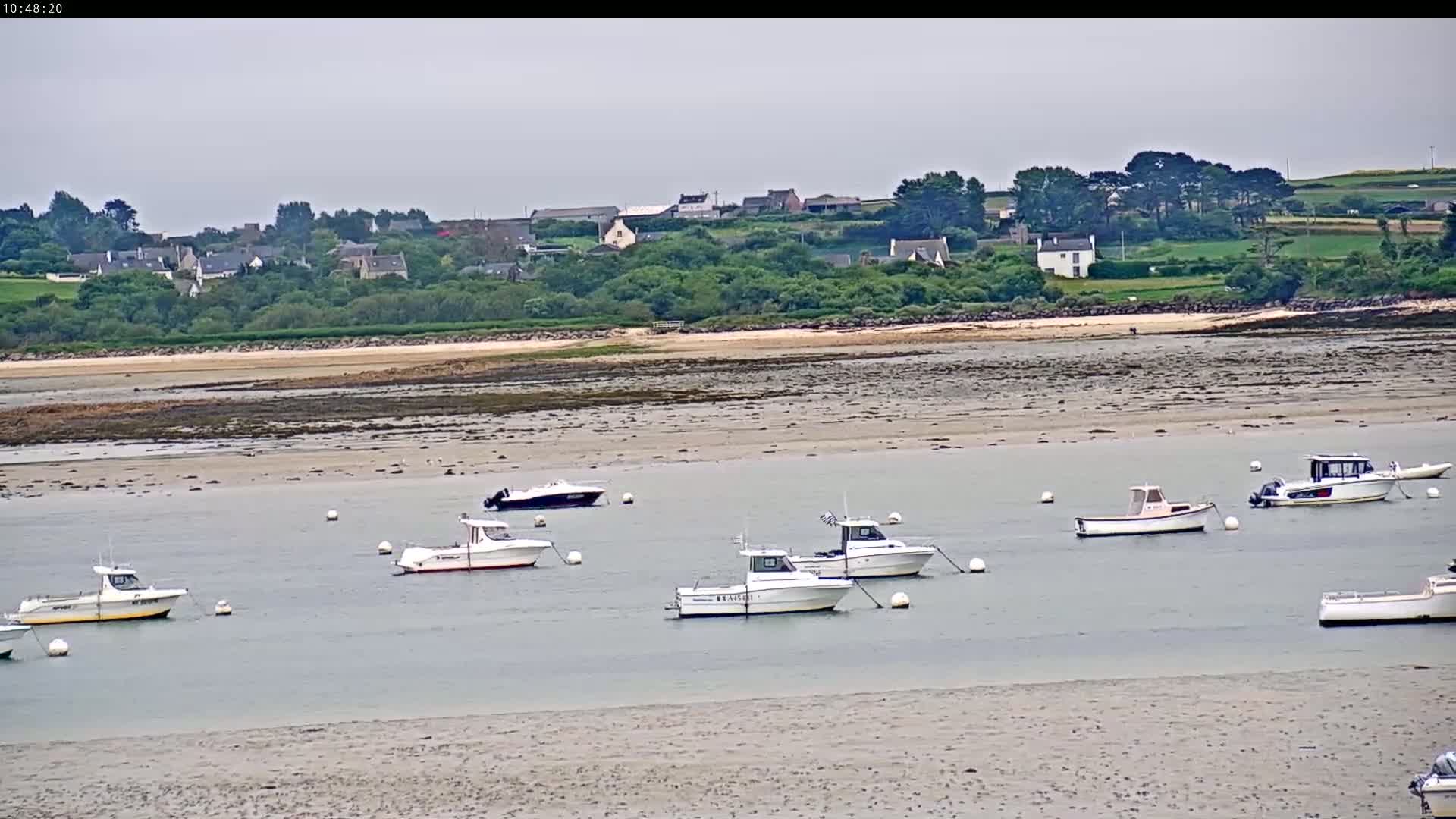 Several small motorboats are moored in calm water near a sandy shore, with a village and green hills visible in the background under an overcast sky.