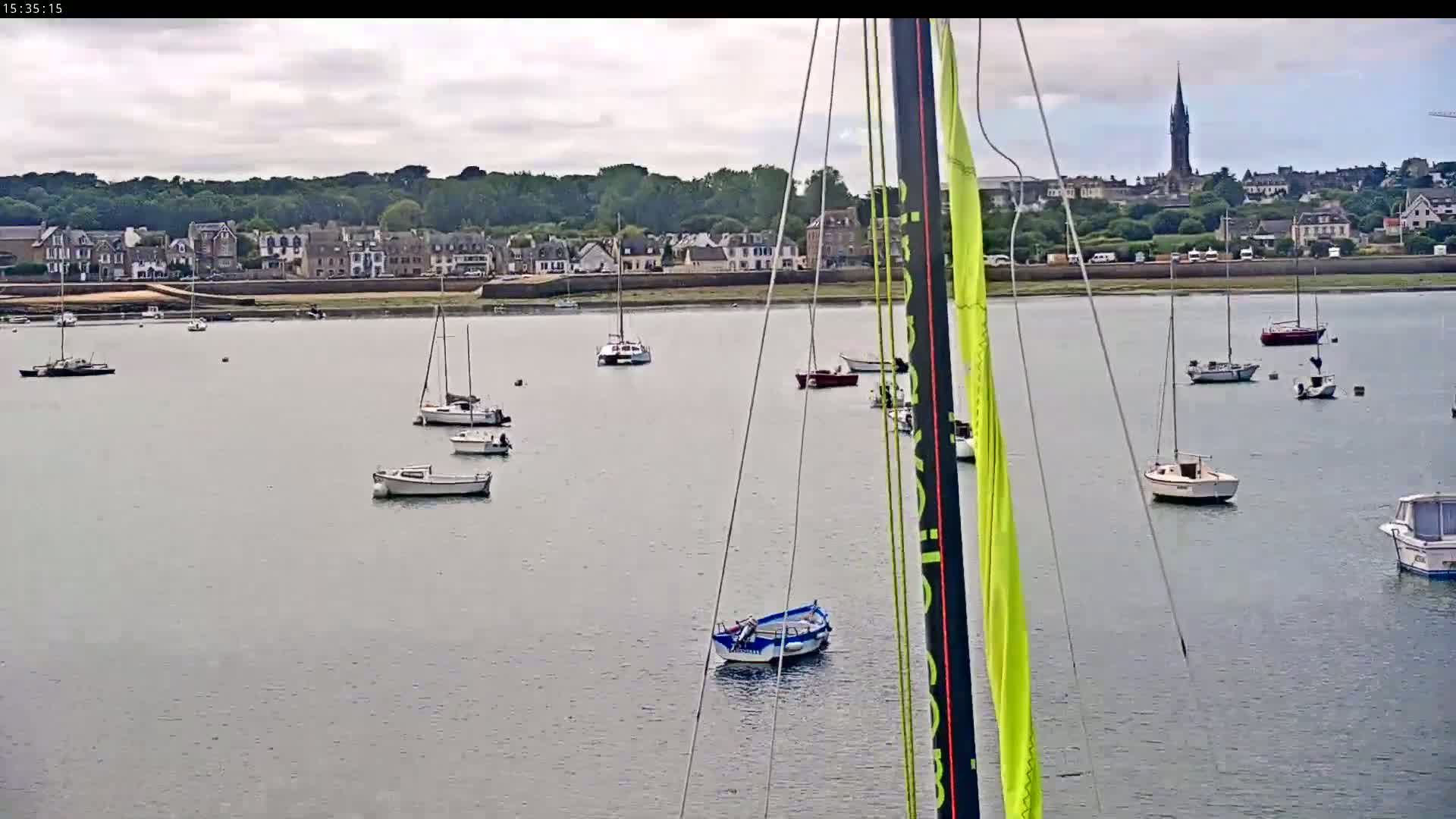 Numerous sailboats and motorboats are moored in a calm bay, with a town and a tall church steeple visible in the background under a cloudy sky.