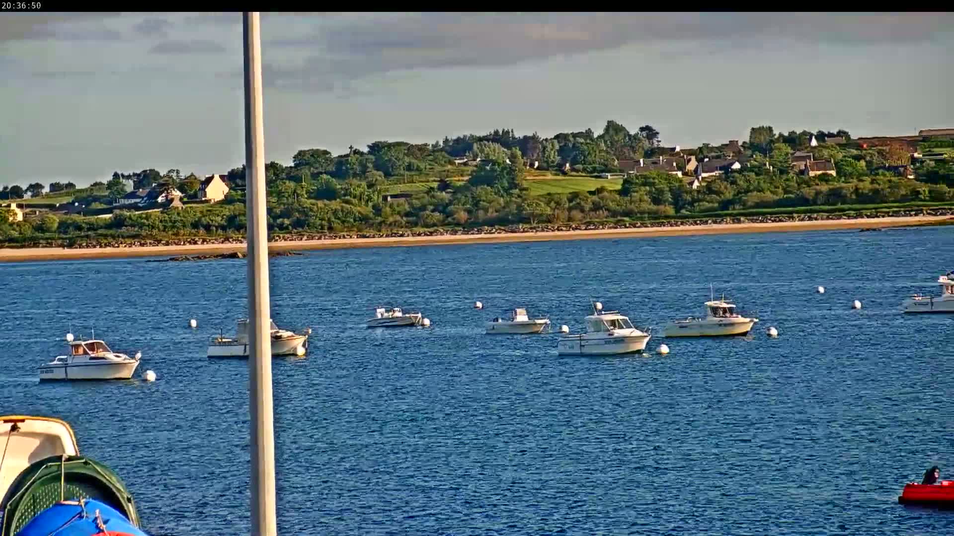 Several motorboats are moored in calm blue water near a sandy shore and a village nestled among green hills under a partly cloudy sky.