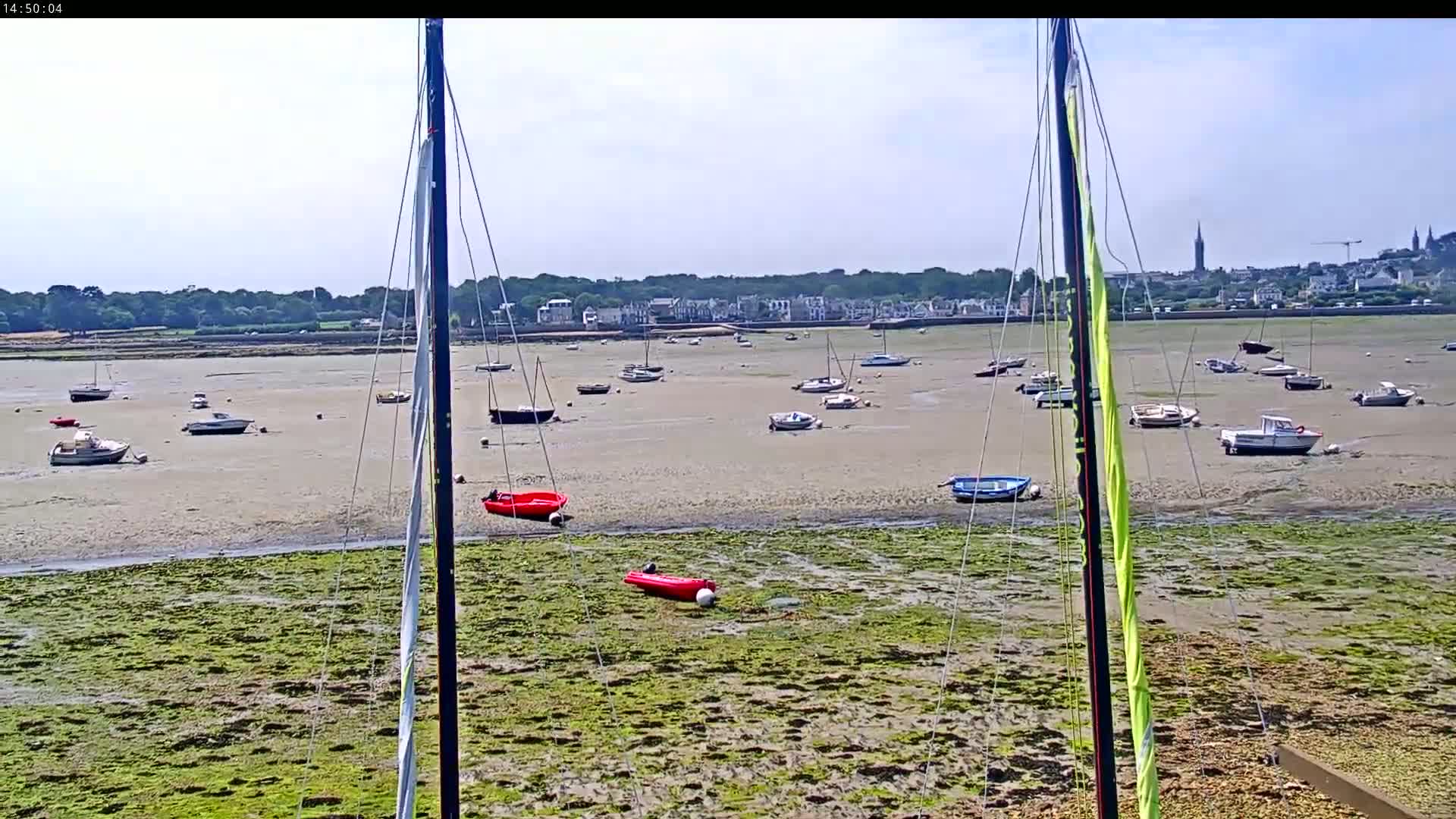 Numerous boats are beached on a tidal flat, with a town visible in the distance on a sunny day.