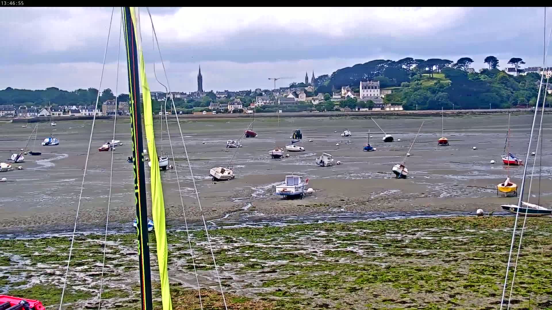 Numerous small boats sit grounded in mudflats at low tide, with a town and green hills visible in the background under a cloudy sky.