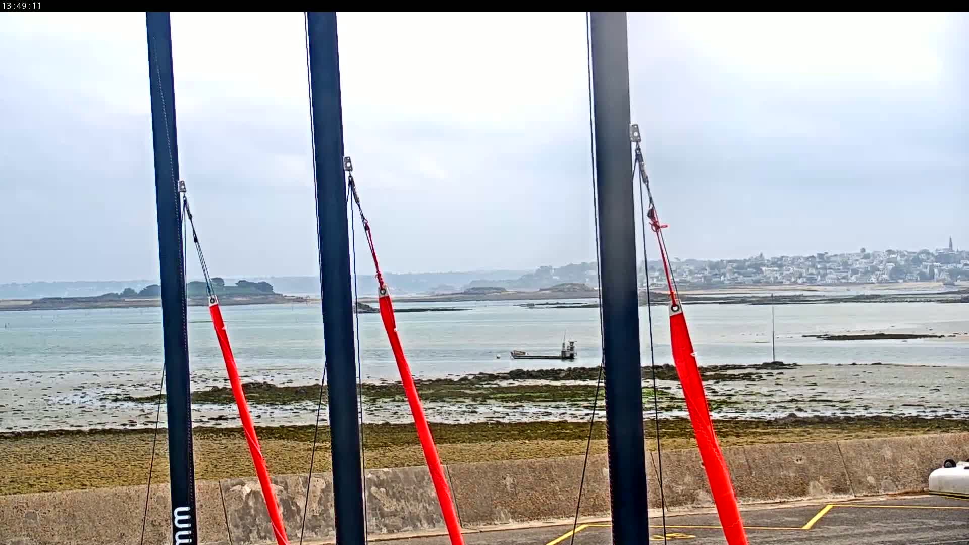 Three dark masts with bright red lines attached are in the foreground, overlooking a calm body of water with exposed mudflats and a distant town under a cloudy sky.