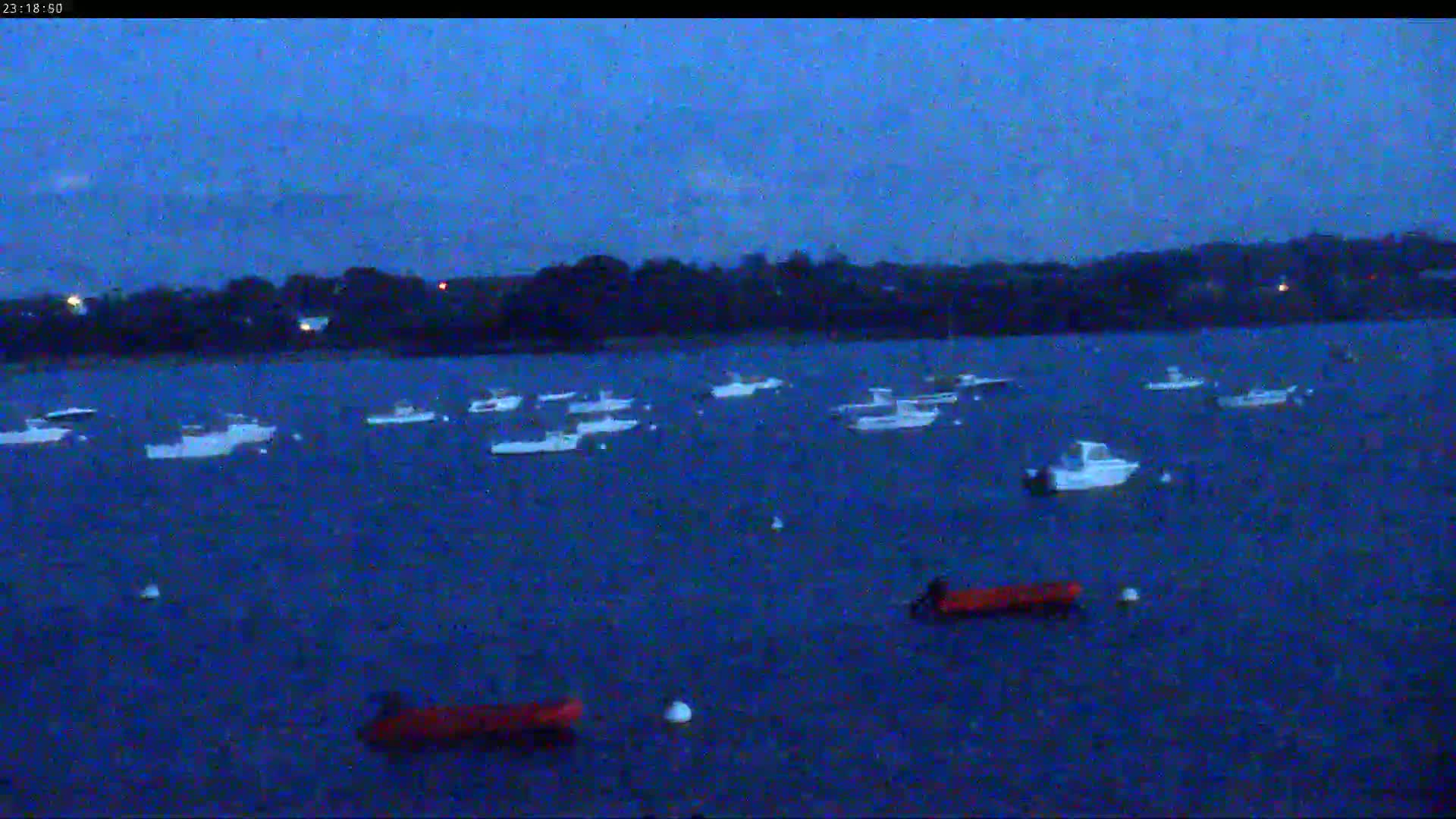 Several sailboats are moored in calm water in front of a town nestled beside a line of trees under a cloudy sky.
