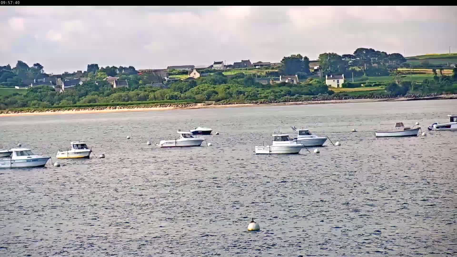 Several small motorboats are moored in calm water off a sandy shore, with a small village nestled amongst green hills in the background under a partly cloudy sky.