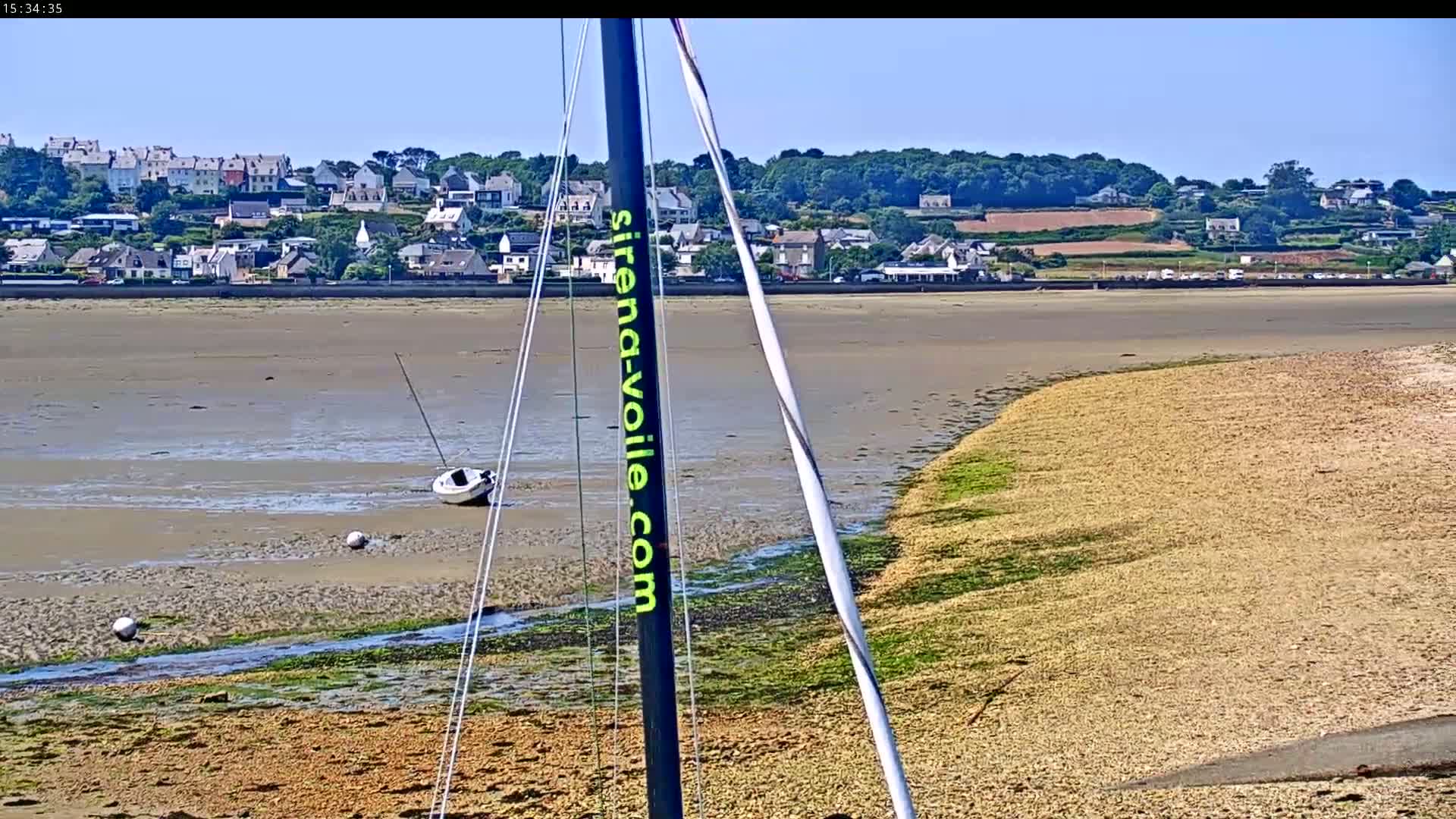 A sailboat mast partially obscures a view of a sandy beach at low tide, with a small sailboat beached nearby and a town visible in the distance under a clear sky.