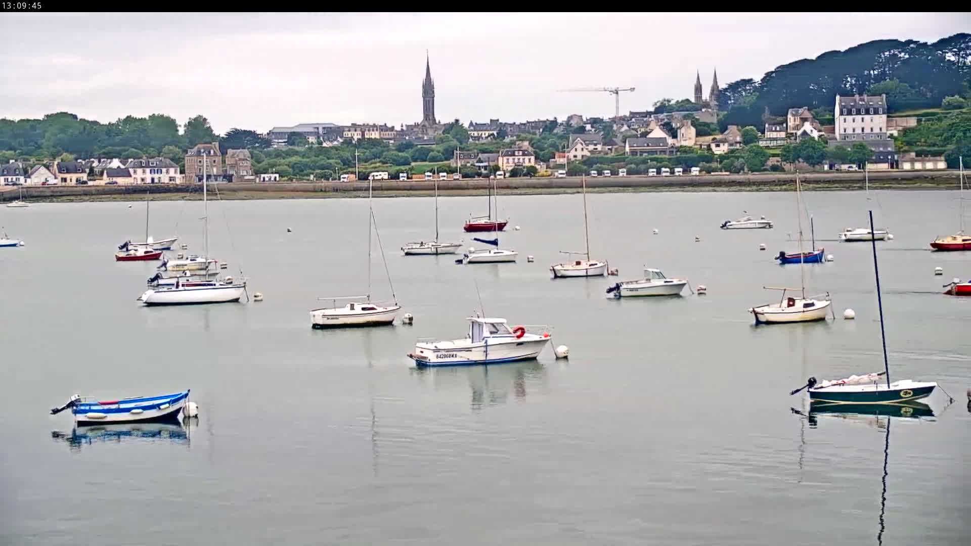 Numerous sailboats and motorboats are moored in a calm harbor under an overcast sky, with a town visible in the background.