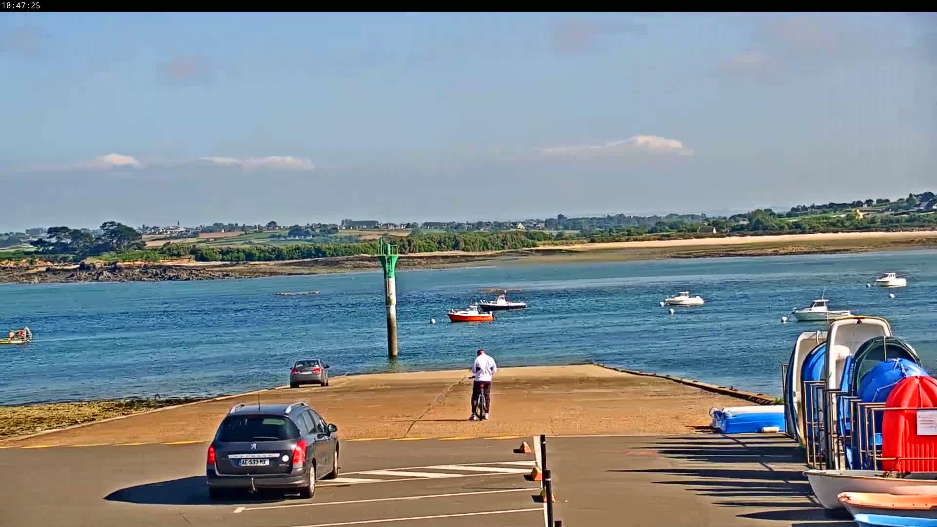 A man stands on a boat ramp overlooking a calm body of water with several boats and cars parked nearby under a mostly sunny sky.