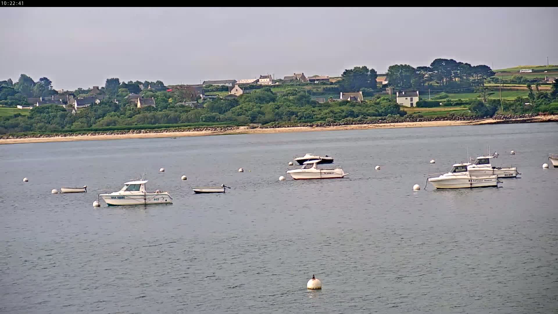 Several motorboats are moored in calm water near a sandy beach and a village on a partly sunny day.