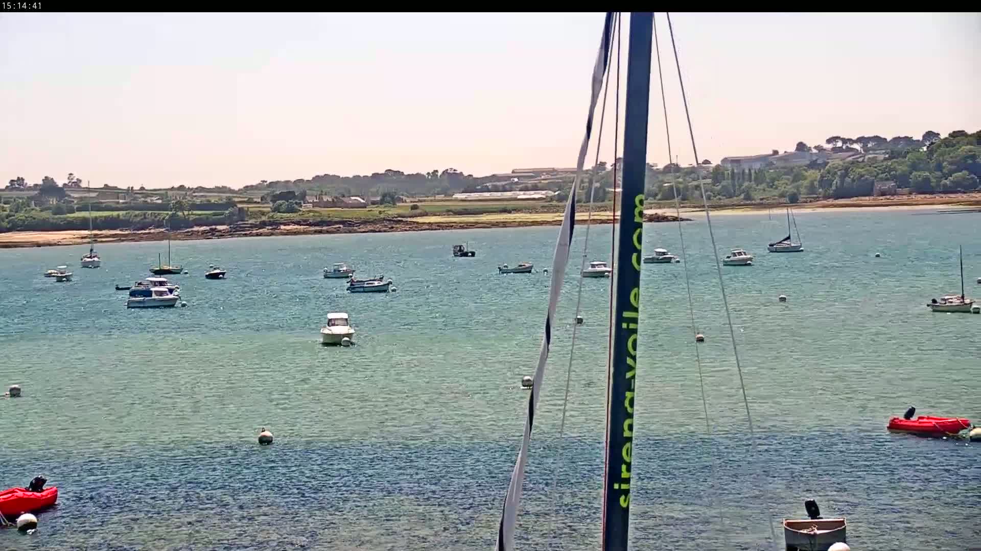 Numerous boats are moored in a calm, sunny bay, with a verdant shoreline in the background.