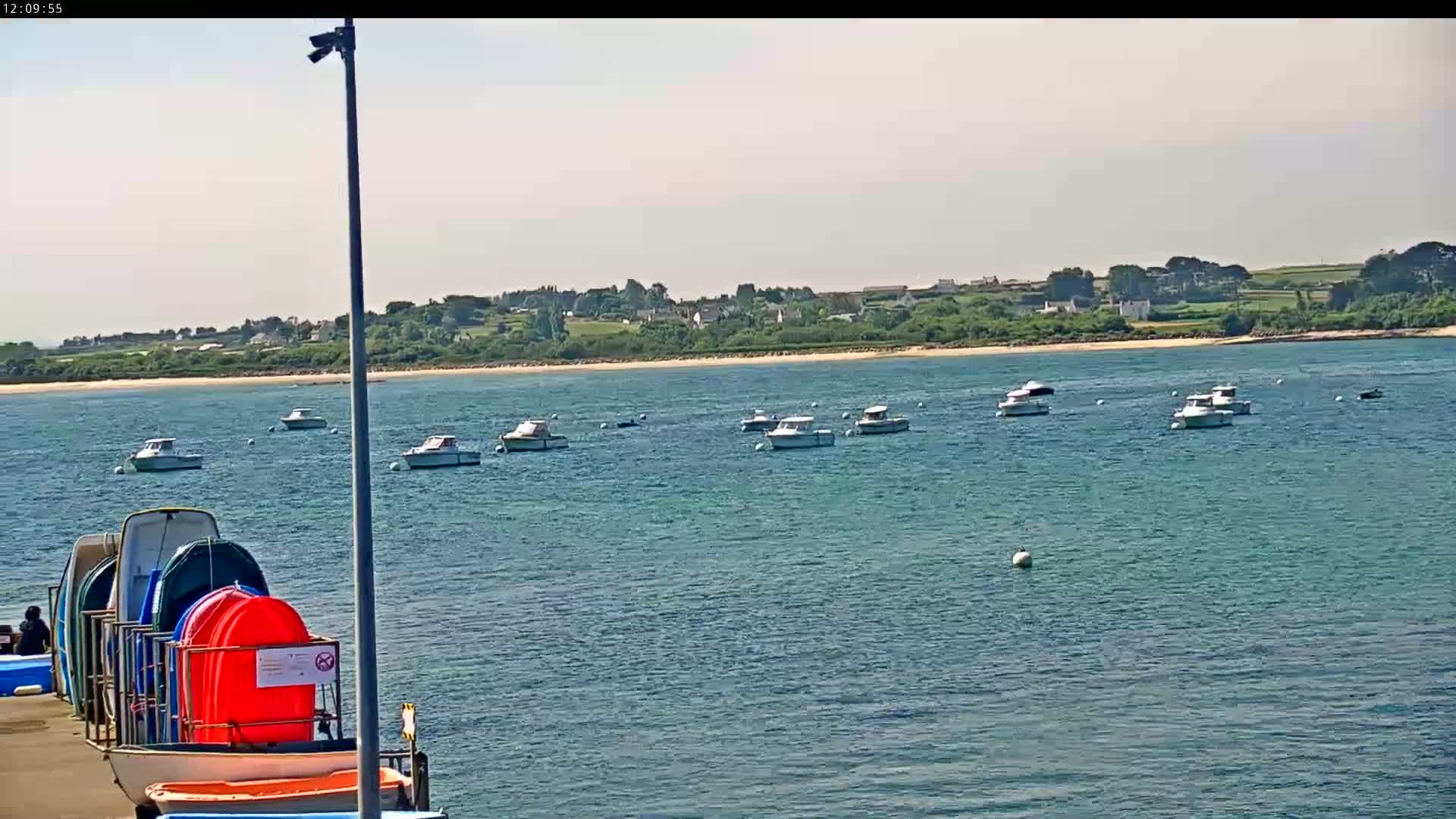 Several motorboats are moored in calm, blue water near a sandy shore and a distant village under a bright, sunny sky.