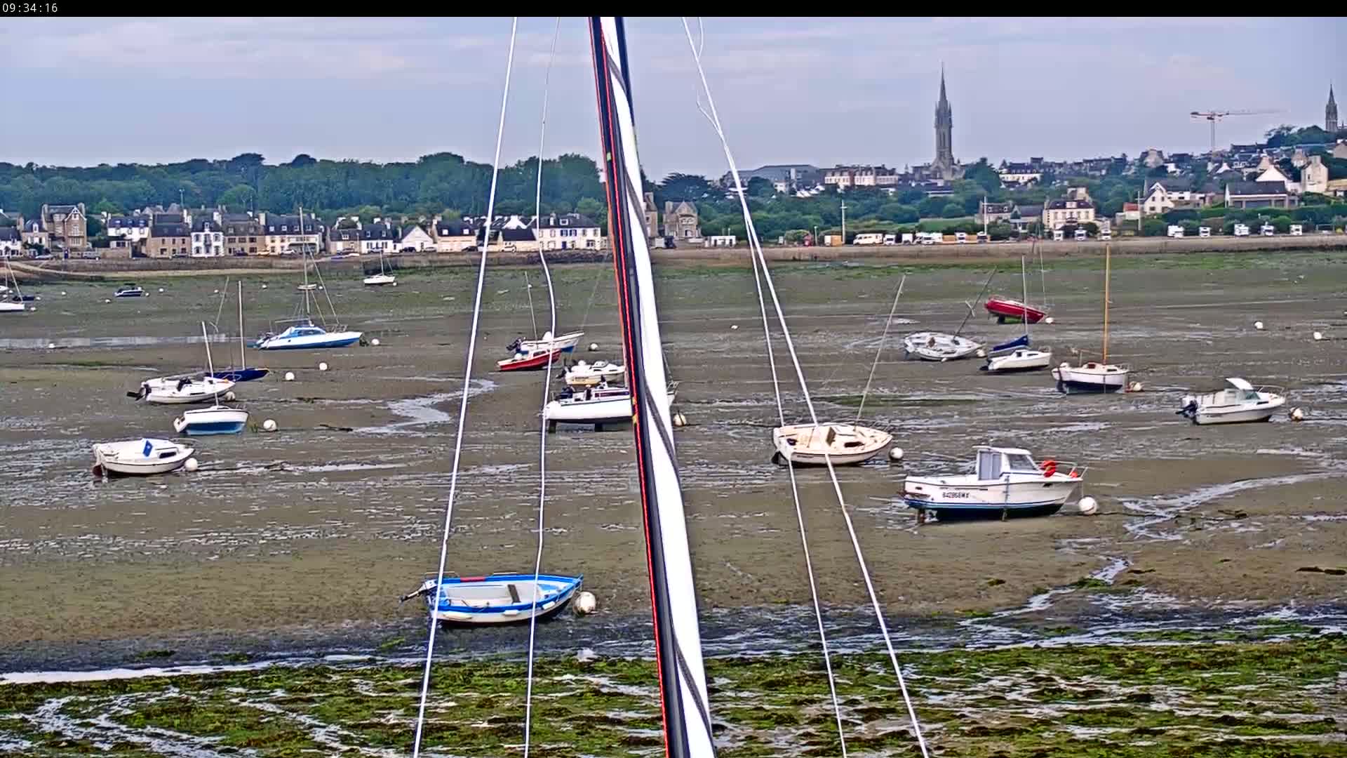 Many sailboats and motorboats are grounded on a muddy tidal flat, with a town and a church spire visible in the background under a partly cloudy sky.