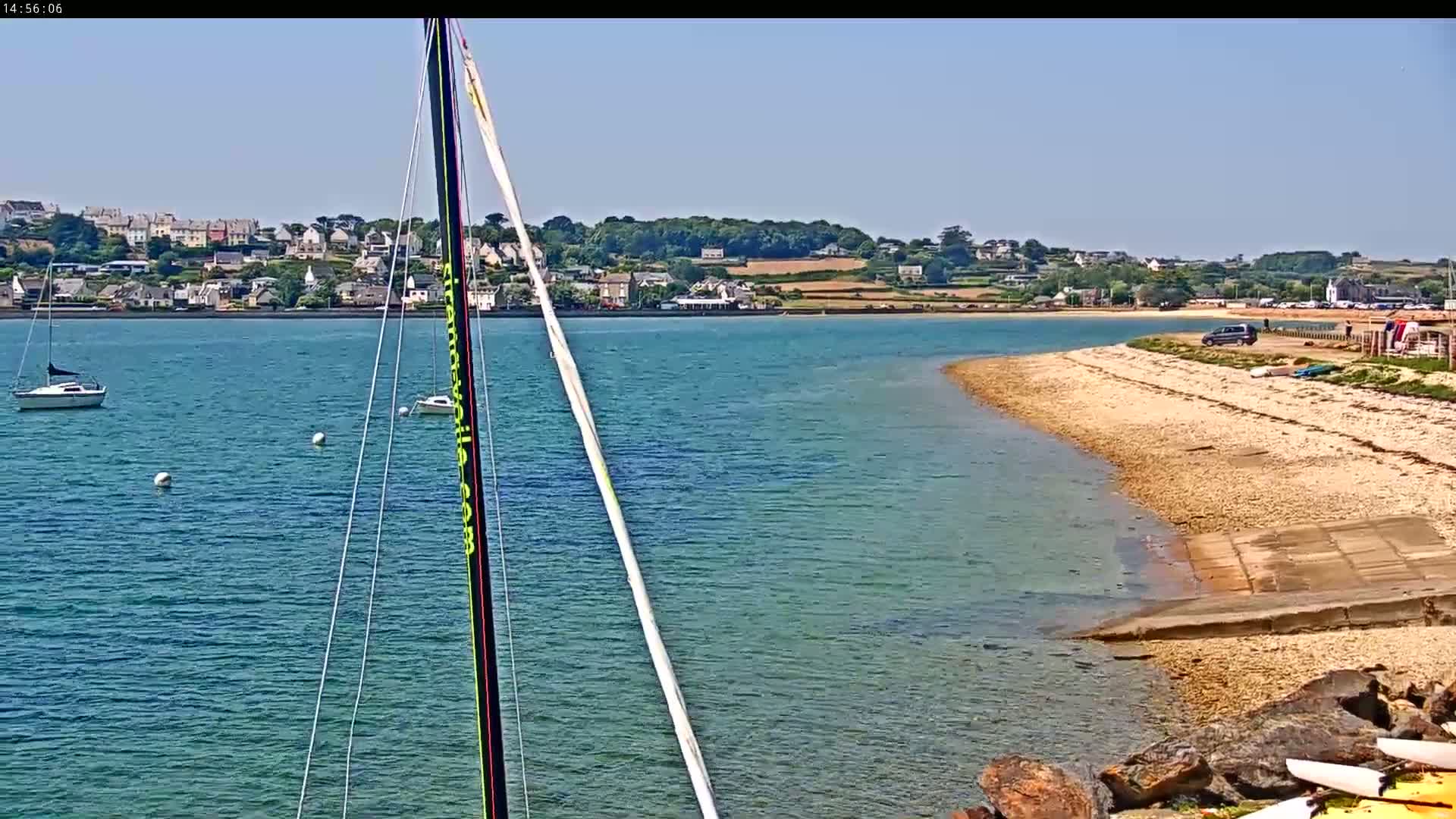 A sailboat mast is in the foreground, overlooking a calm, blue body of water with a sandy shoreline and distant buildings under a clear, sunny sky.