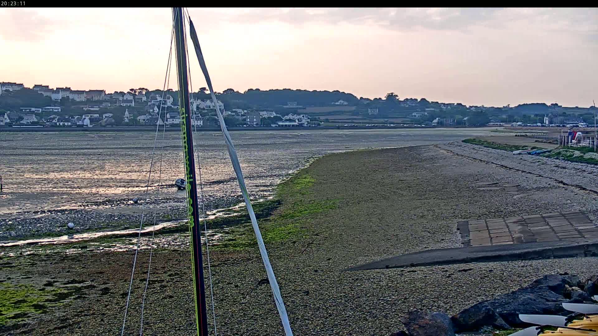 A mostly cloudy sunset over a calm body of water, showing a long, pebble beach leading to a small town on the far shore, with the mast of a sailboat in the foreground.