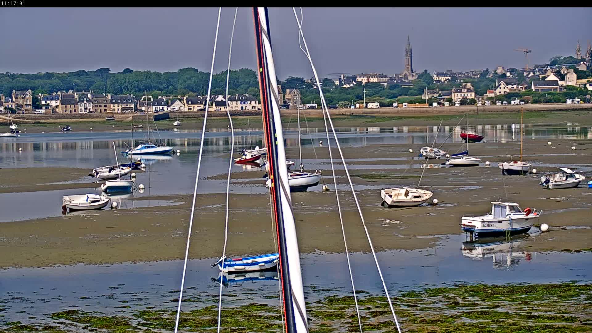 Numerous small boats sit in shallow, muddy water at low tide near a town with buildings and a church steeple in the background on a sunny day.