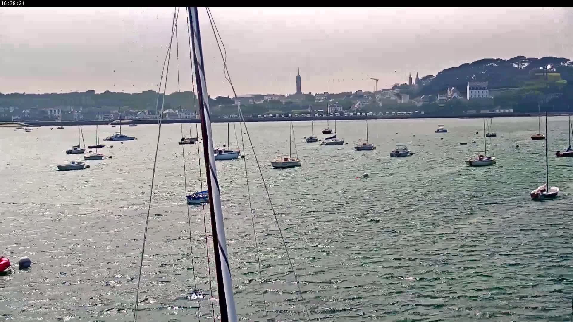 Numerous sailboats are anchored in a calm body of water in front of a distant town on a partly sunny day.