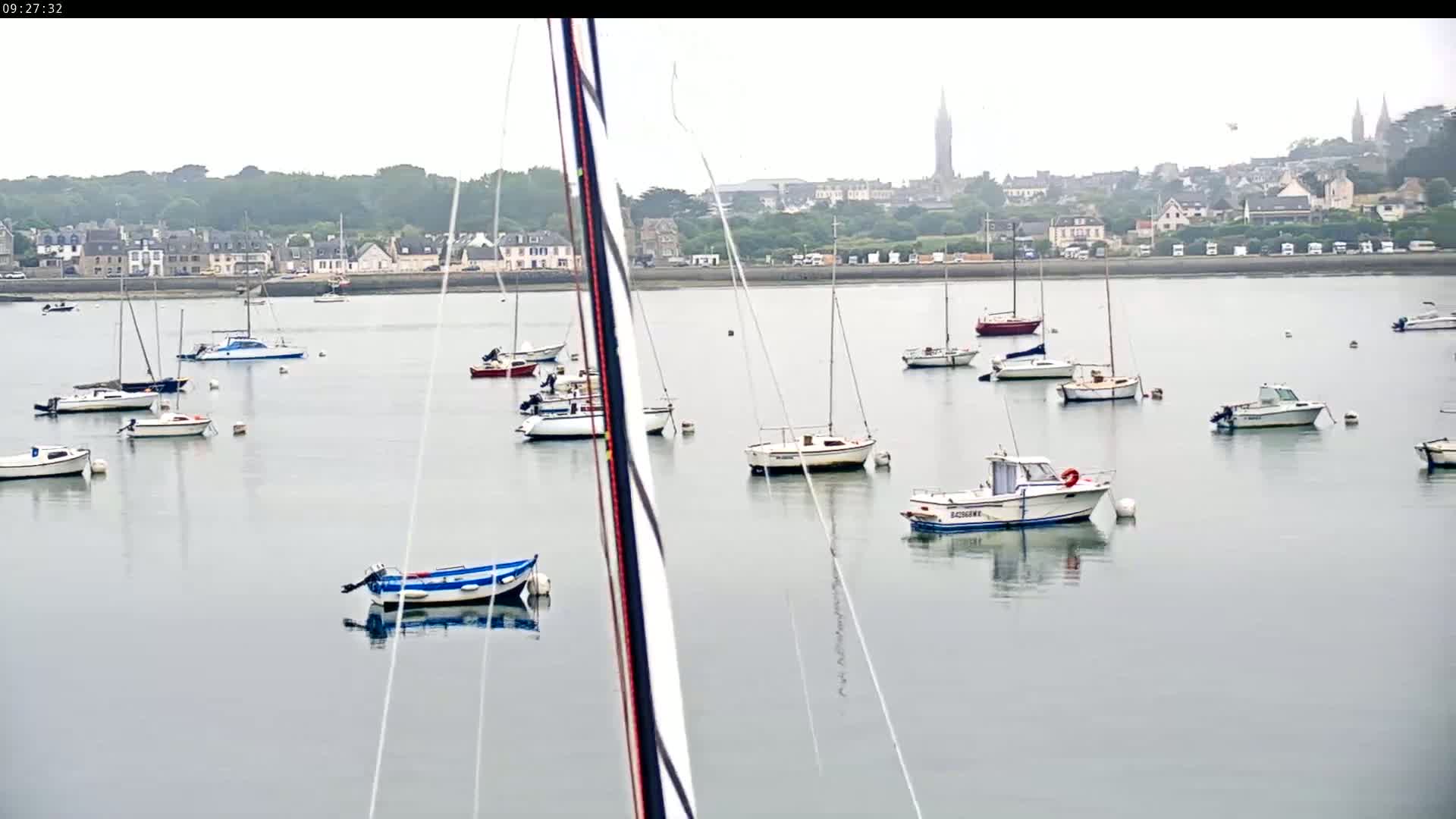 Numerous sailboats and motorboats are moored in calm, overcast water in front of a distant town.