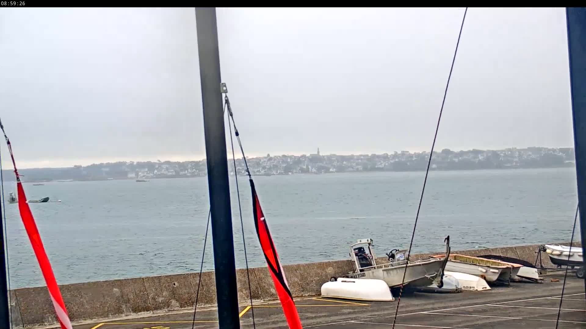 Several small boats are beached on a paved area next to a body of water under an overcast sky, with a town visible in the distance.