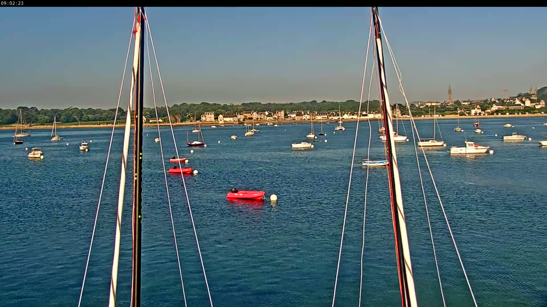 From a sailboat's mast, numerous boats are anchored in calm, blue water, with a distant town visible under a clear sky.