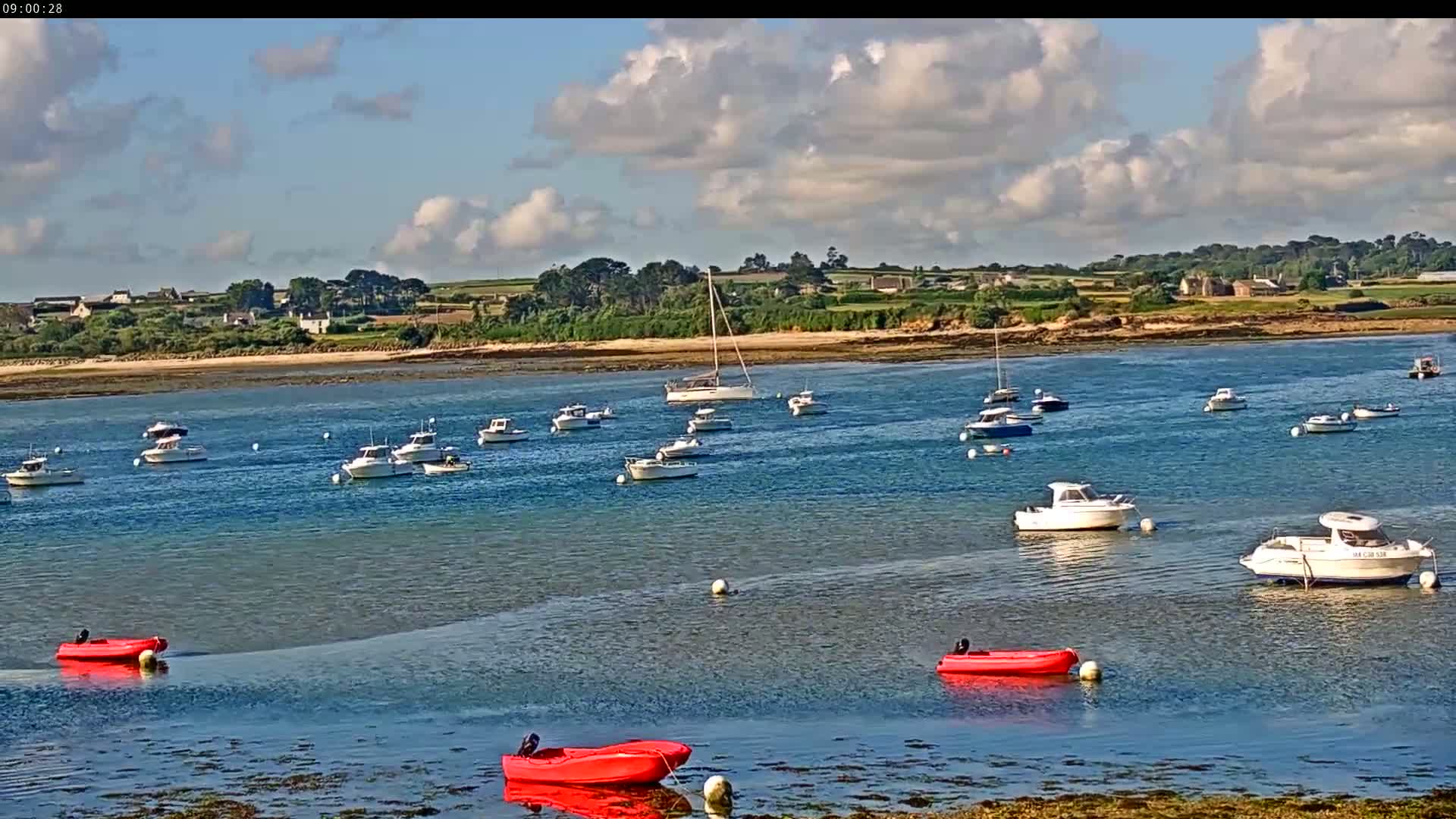 Numerous boats are moored in a calm bay under a partly cloudy sky, with a sandy shoreline and distant land visible in the background.