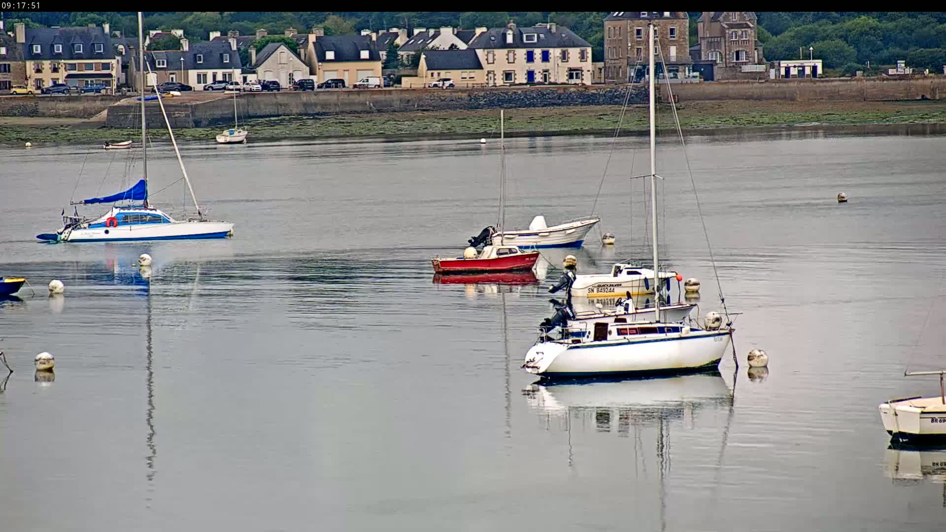 Several sailboats and motorboats are moored in calm water in front of a stone waterfront village on an overcast day.