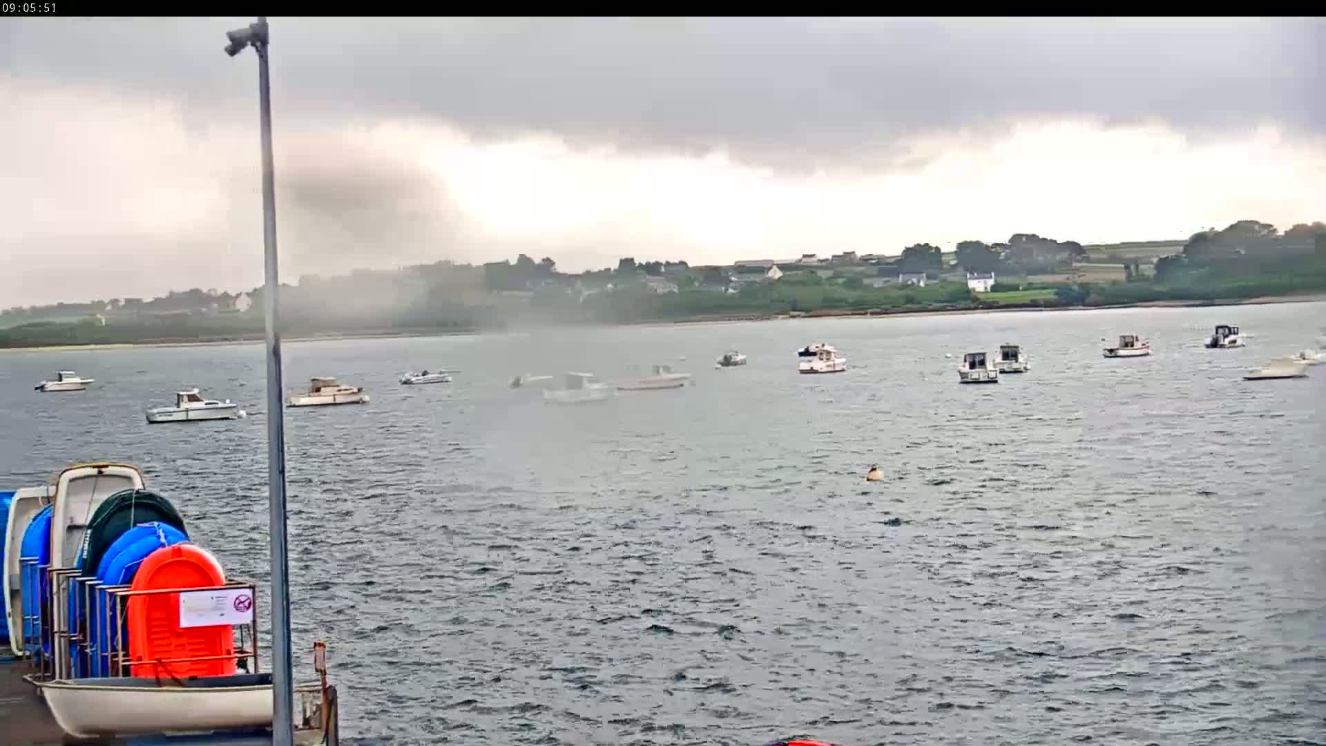 Numerous boats are moored on a calm body of water under an overcast sky, with a hazy mist near the shoreline and stacked colorful small boats visible in the foreground.