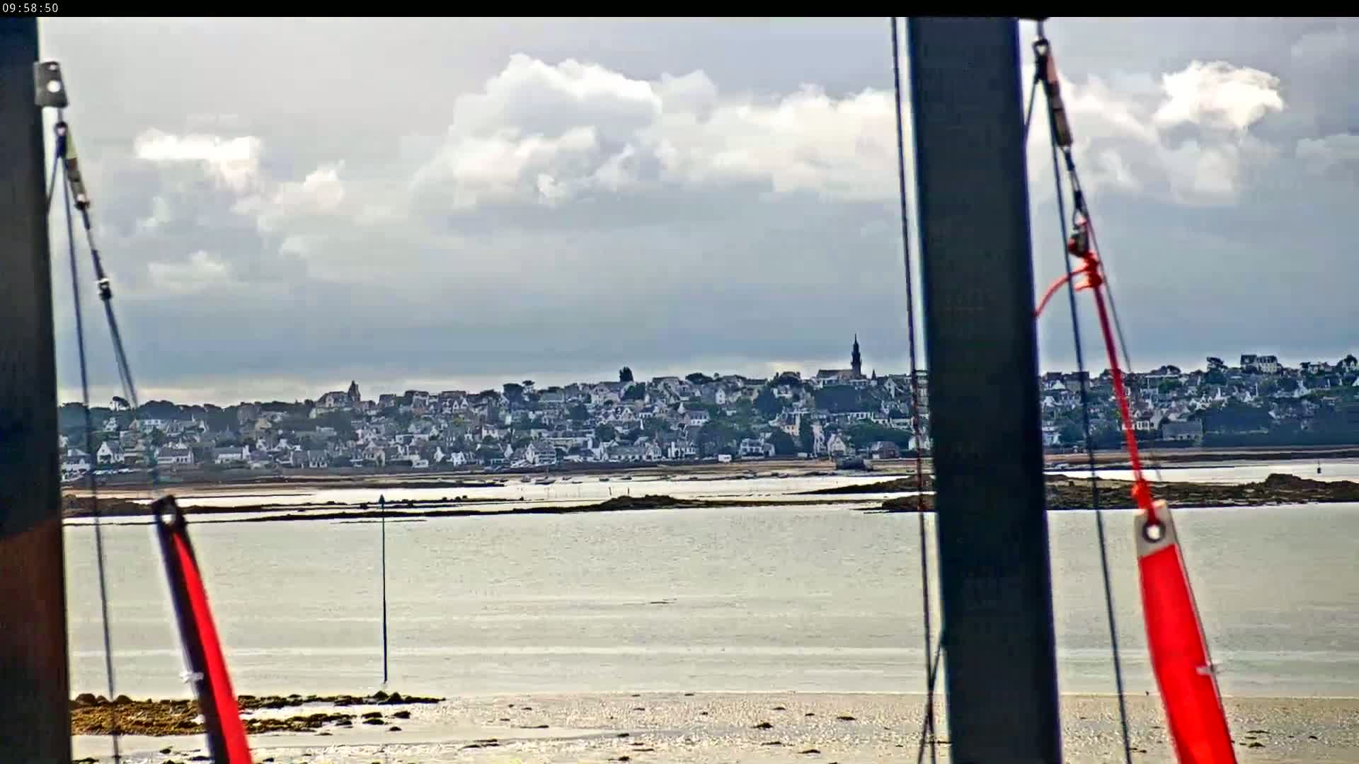 A coastal town is visible across a calm body of water under a cloudy sky, viewed from behind what appears to be the mast of a sailboat.