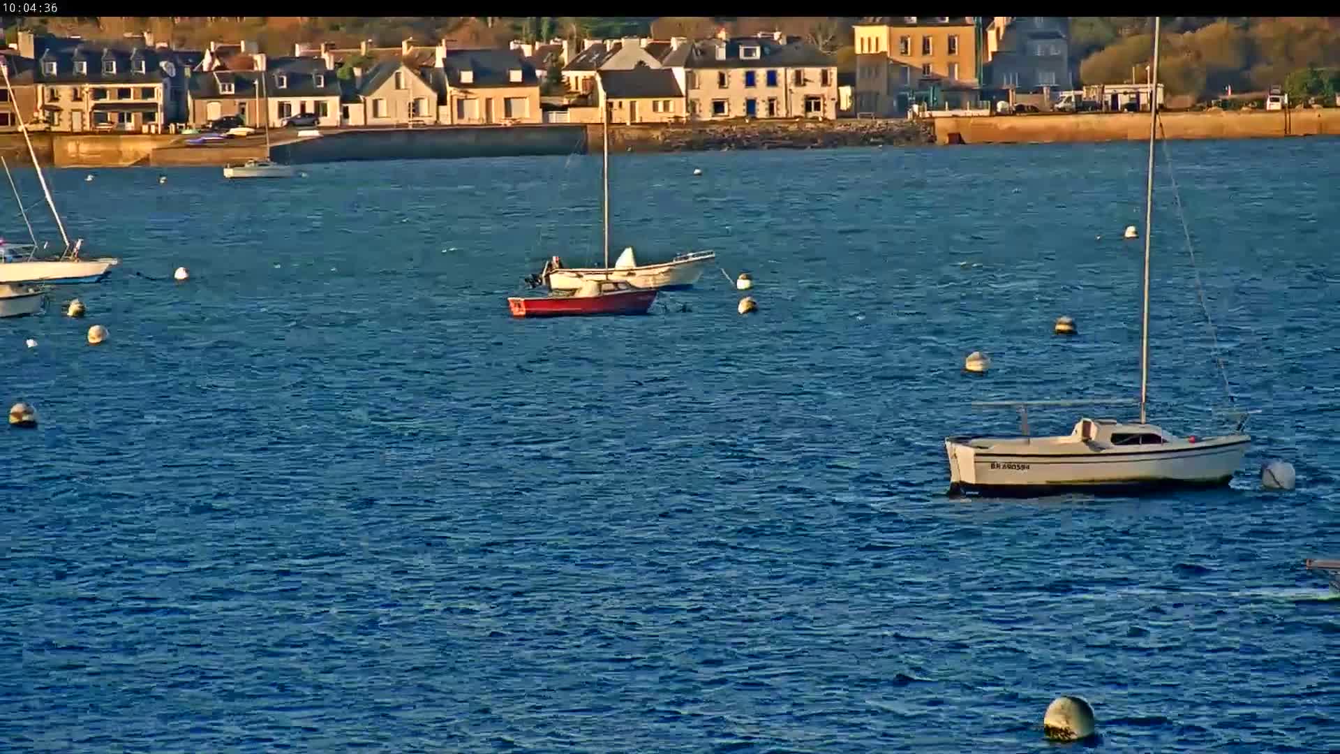 On a clear and sunny day, several boats are moored in a bay with rippling blue water, backed by a picturesque coastal town.