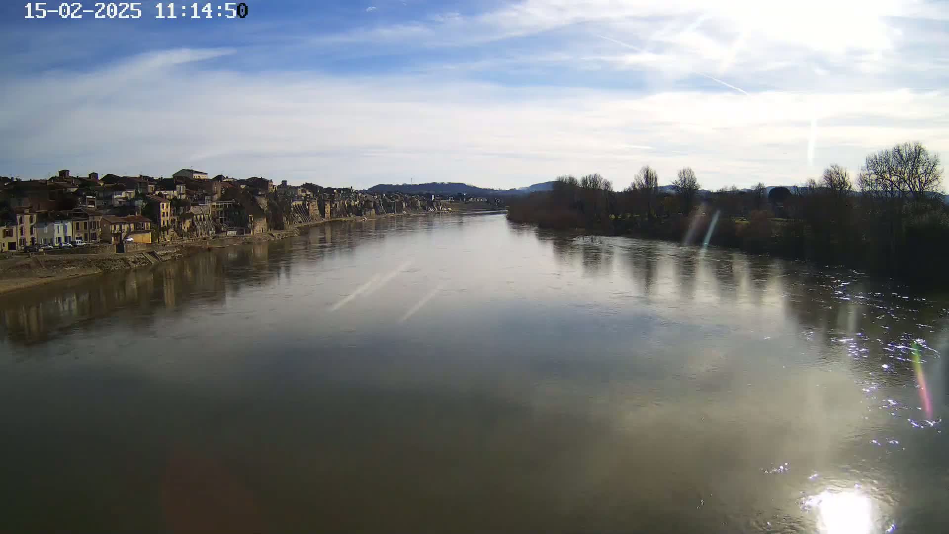 A wide, calm river reflects a partly sunny sky and the buildings and trees on its banks.