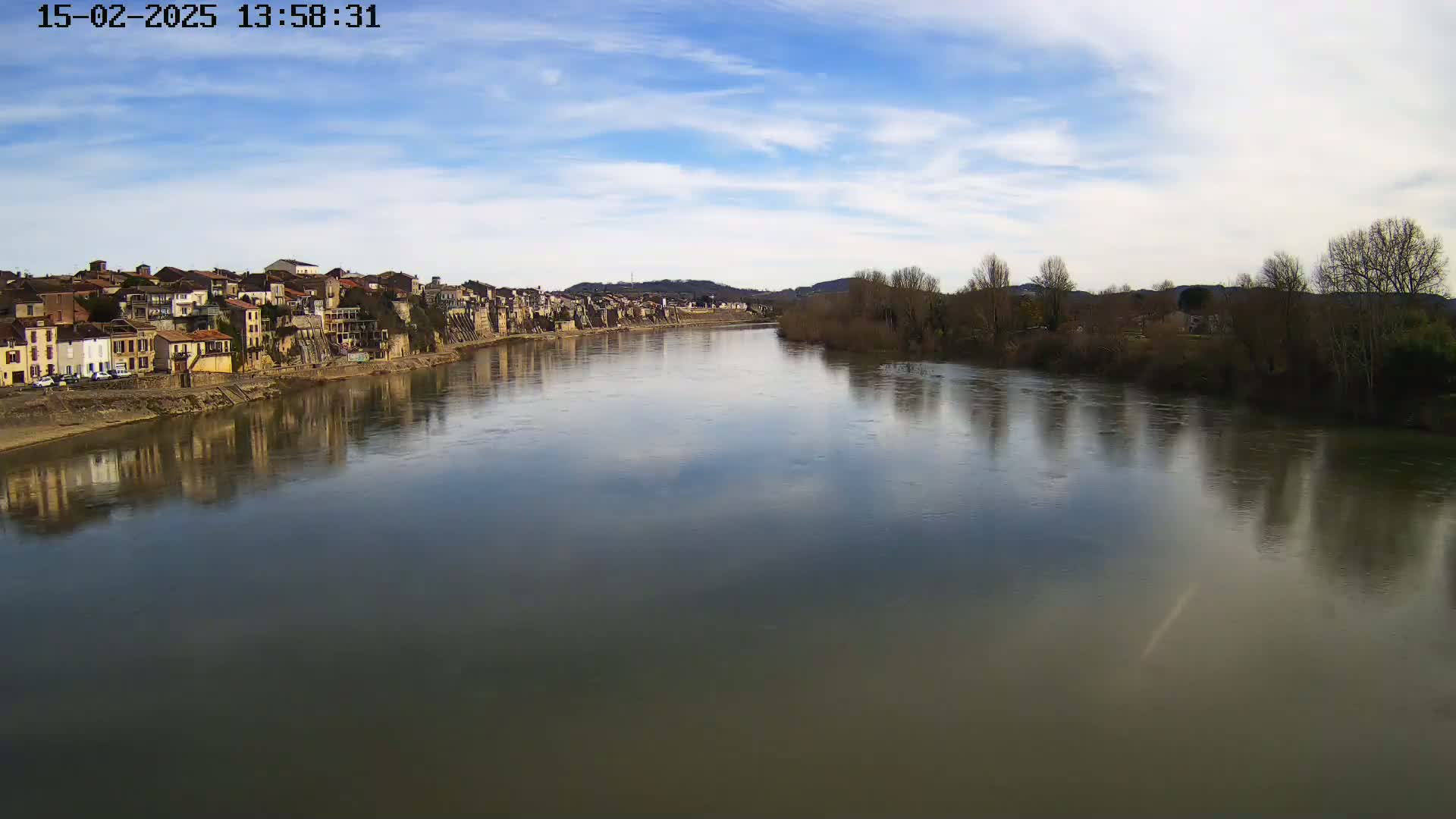 A calm river reflects a partly cloudy sky and the buildings of a town on its banks.