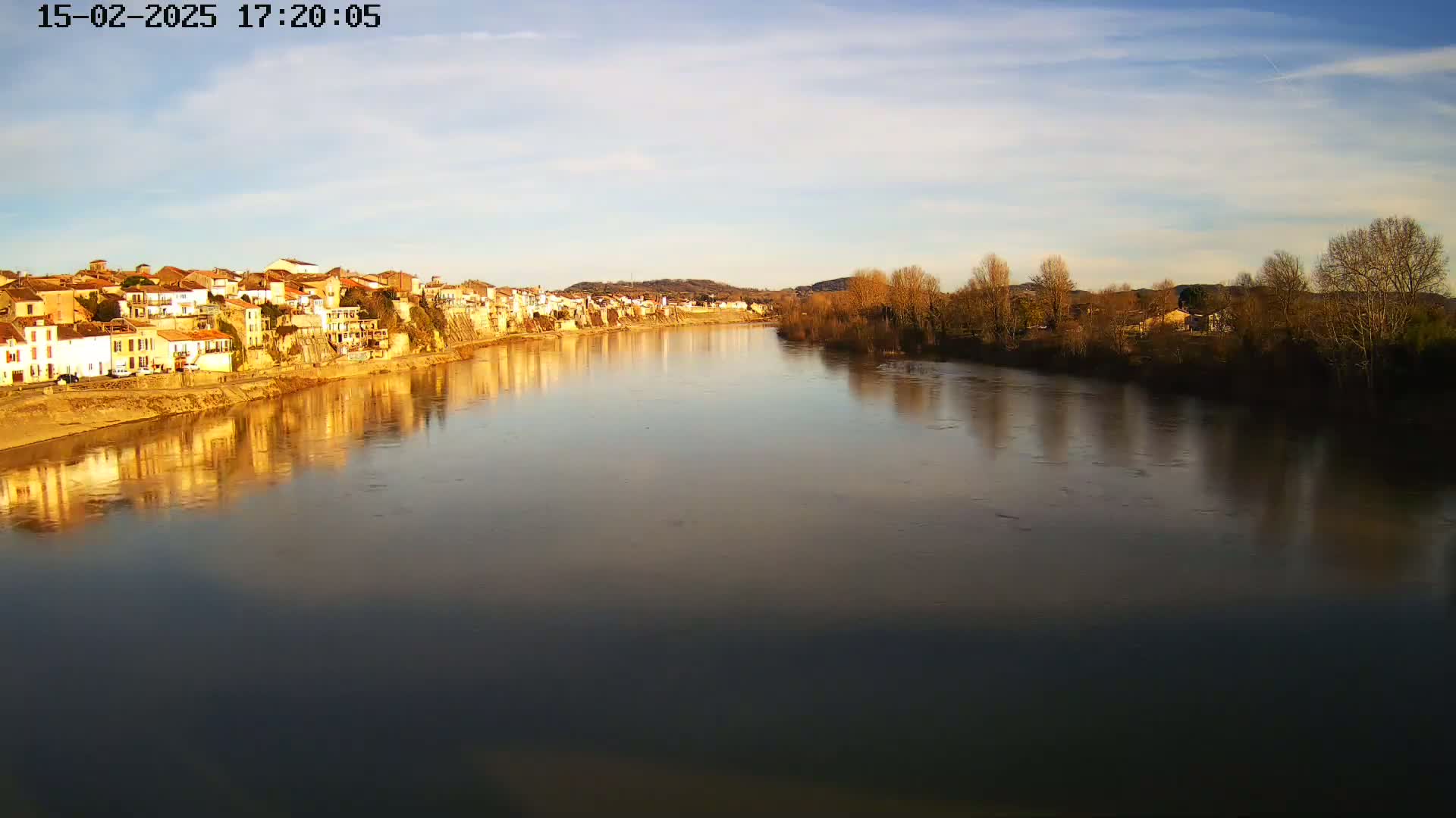 A calm river reflects the golden light of a clear, late afternoon sky and a row of buildings on its banks, lined with leafless trees.