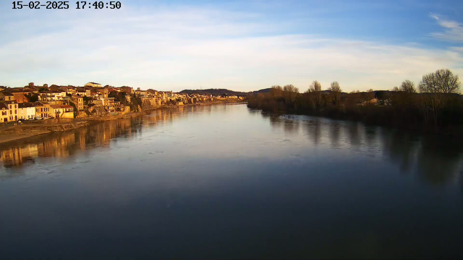 A calm river reflects a town of tan buildings under a mostly clear, light blue sky.