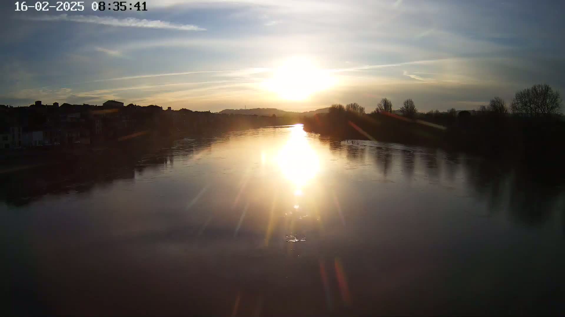 A calm river reflects the bright sunset, with silhouetted trees and buildings lining its banks under a mostly clear sky.