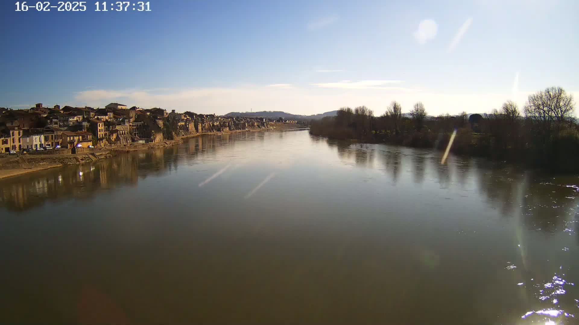 A calm river reflects a town and bare winter trees under a mostly clear, bright sky.