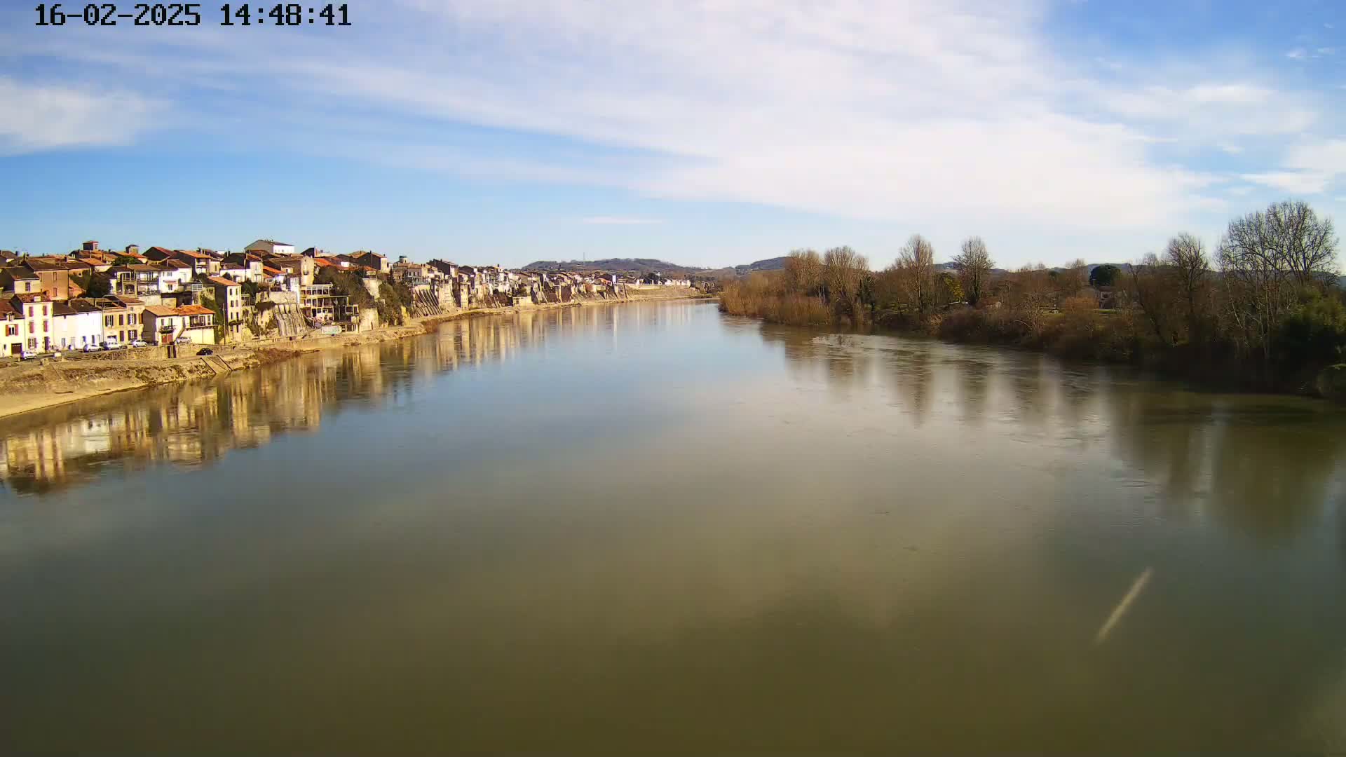 A river reflects a town of closely-packed buildings under a mostly sunny sky.