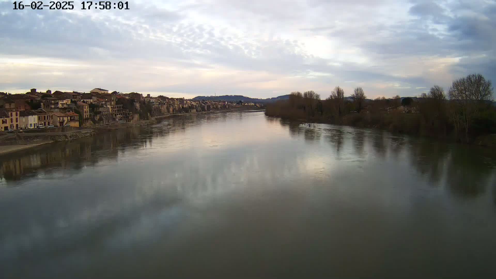 A wide river reflects a cloudy, dusky sky as it flows past a town with buildings lining its bank.
