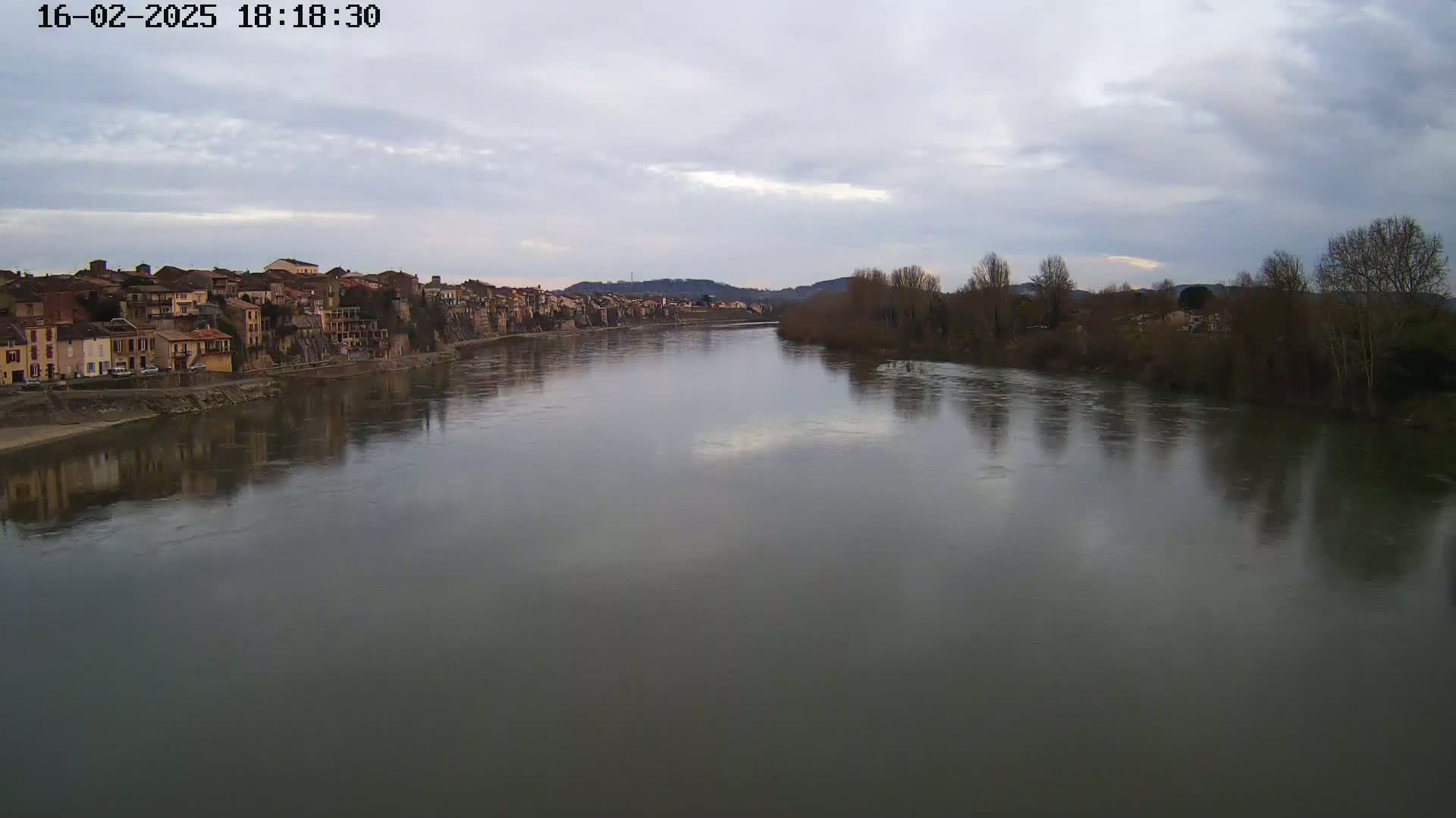 A wide river reflects a cloudy sky and the silhouettes of leafless trees and a town on its banks.