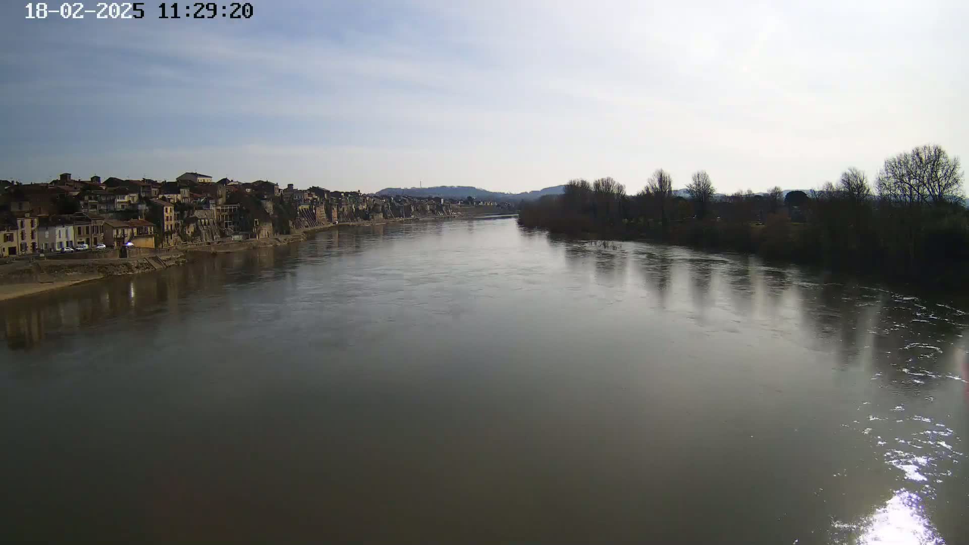 A wide, calm river reflects a mostly cloudy sky and the silhouettes of leafless trees on its banks, with a town of low buildings visible on the opposite shore.