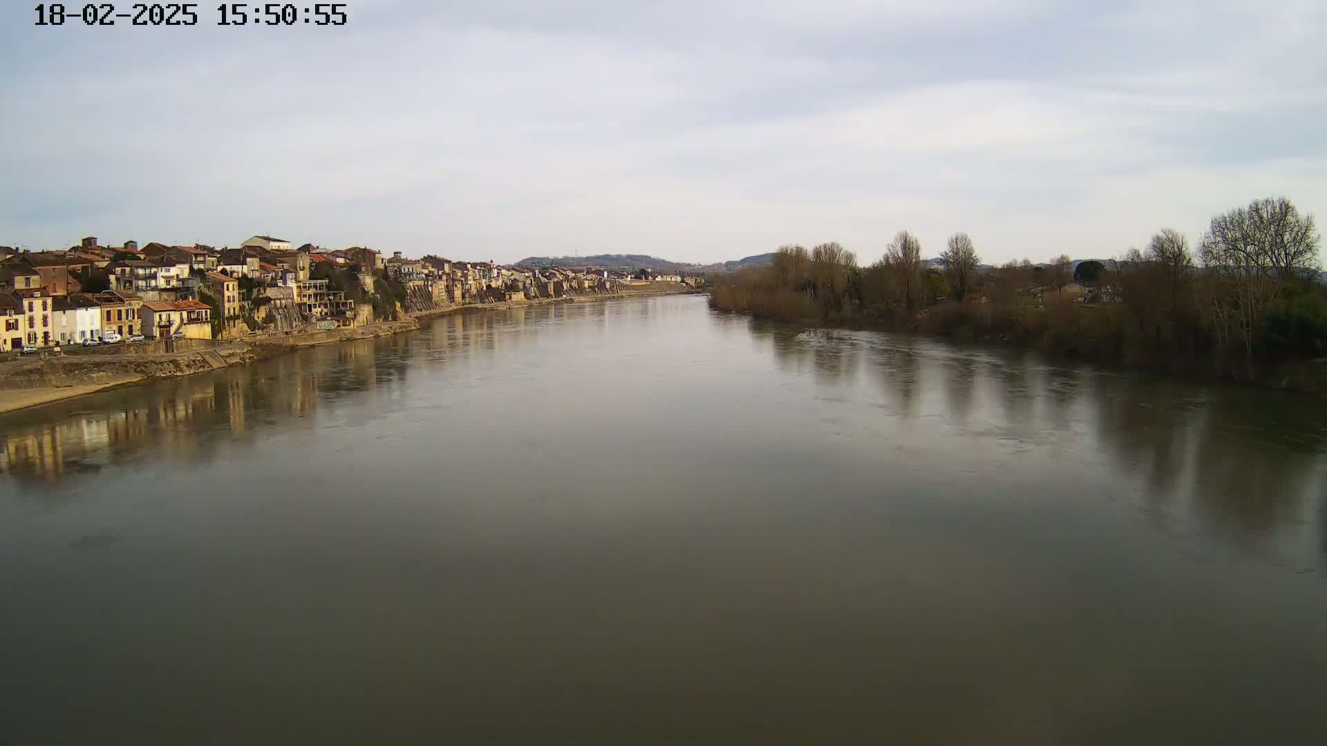 A wide, calm river reflects a town of closely-packed buildings on one bank and bare winter trees on the other under a cloudy sky.