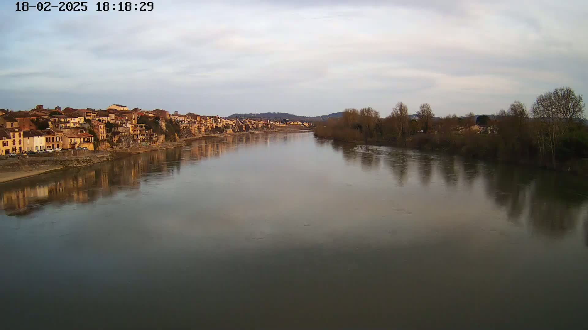 A calm river reflects a row of buildings on one bank and bare trees on the other under a cloudy sky.
