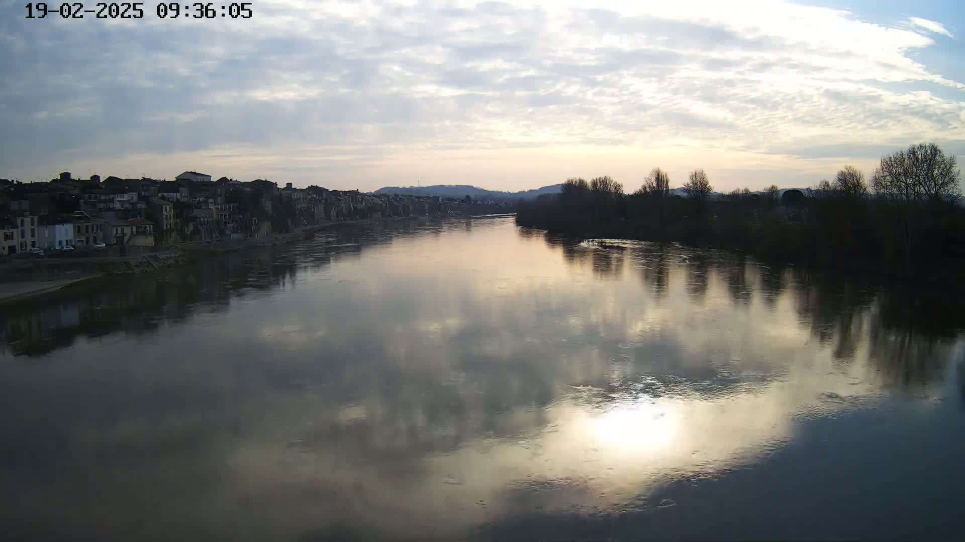 A calm river reflects a cloudy sunset sky, with a town on one bank and bare trees lining the opposite shore.