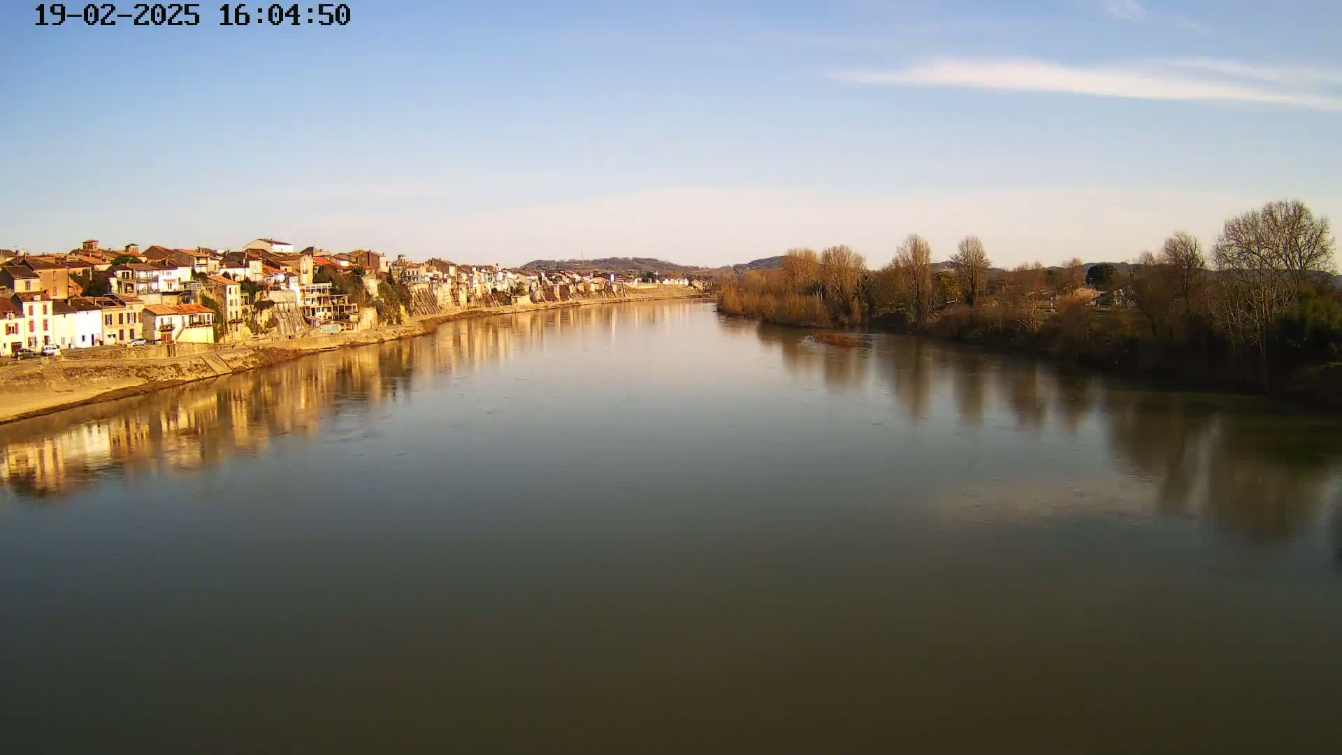A calm river reflects a row of buildings and bare winter trees under a clear, light blue sky.