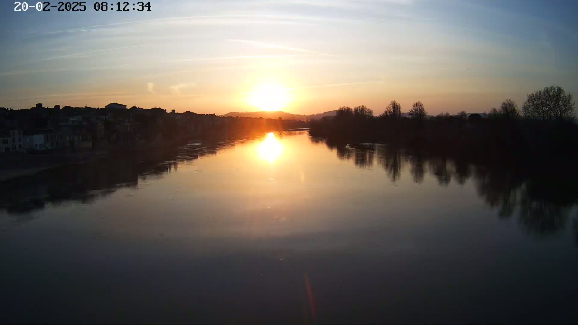A calm river reflects a setting sun, with buildings and trees silhouetted against the warm-colored sky.