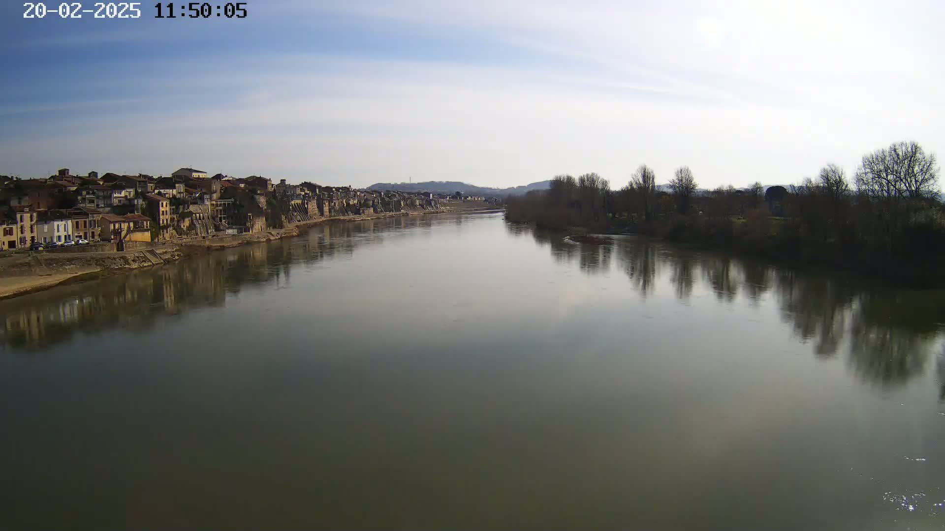 A calm river reflects a town and bare trees under a partly cloudy sky.
