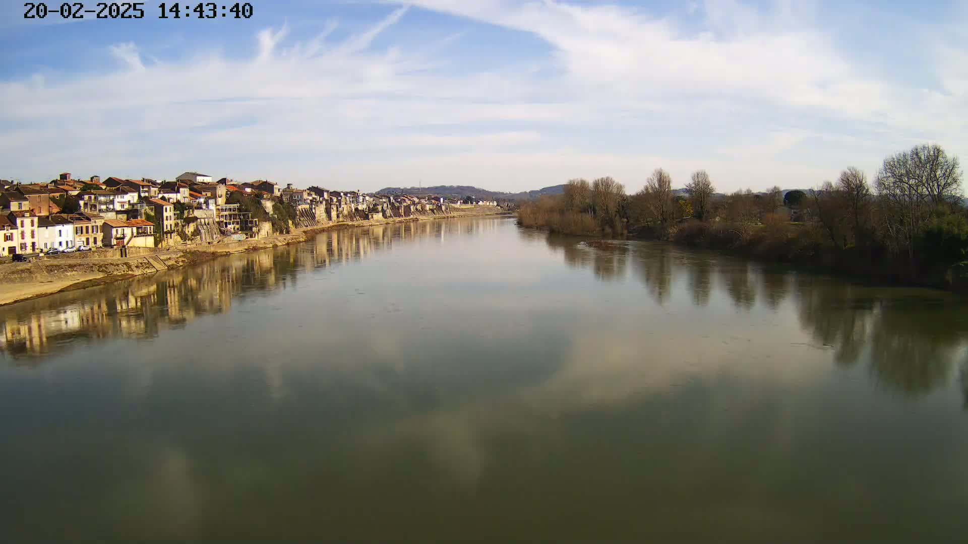 A calm river reflects a row of buildings and bare winter trees under a partly cloudy sky.