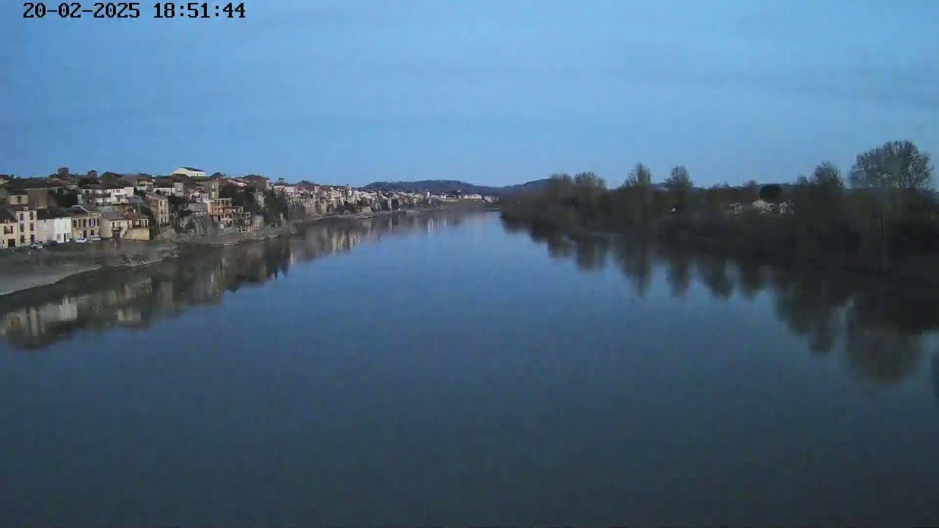A calm river reflects a town's buildings and bare winter trees under a twilight sky.