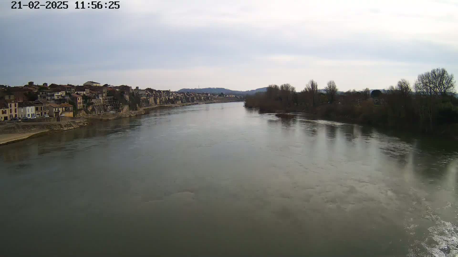 A wide, calm river flows past a town of buildings with brown roofs, under a mostly cloudy sky.