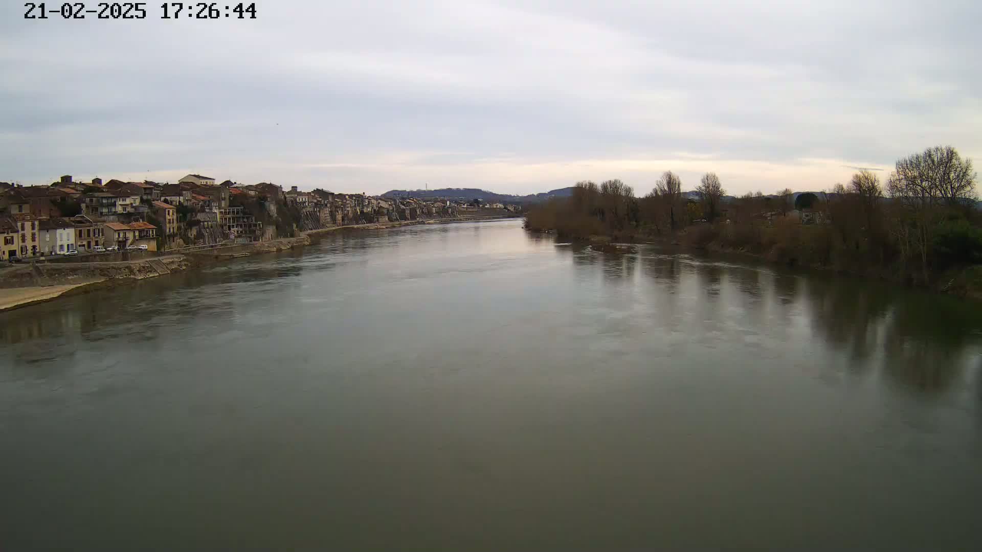 A wide, calm river flows past a row of buildings on one bank and bare trees on the other under an overcast sky.